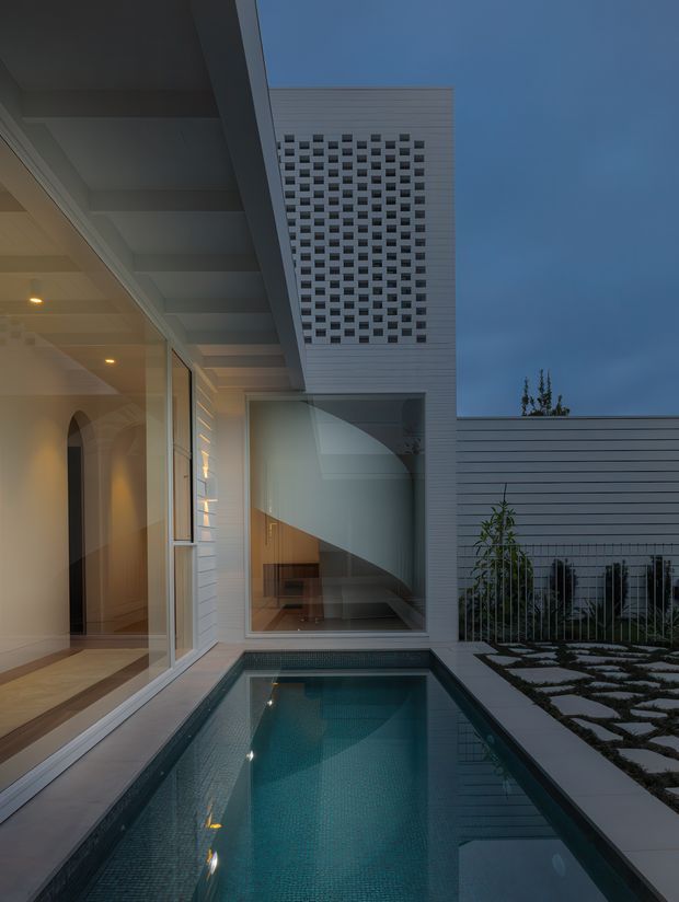 Dusk view of a refined modern courtyard centered on a narrow rectangular swimming pool. White horizontal cladding, extensive floor-to-ceiling glazing, a covered walkway, and a perforated breeze-block-style upper screen create a crisp geometric facade. Warm interior lighting contrasts with the cool blue evening sky, while a small planted gravel garden sits beside the pool.