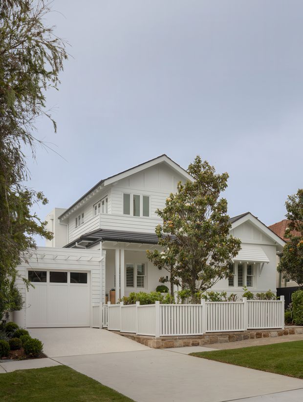Exterior street view of a light-painted, gable-roofed two-storey suburban home with white weatherboard cladding, traditional trim, shuttered windows, a covered entry, attached single garage, front fence, paved driveway, and landscaped garden. The composition is focused on the residential facade.
