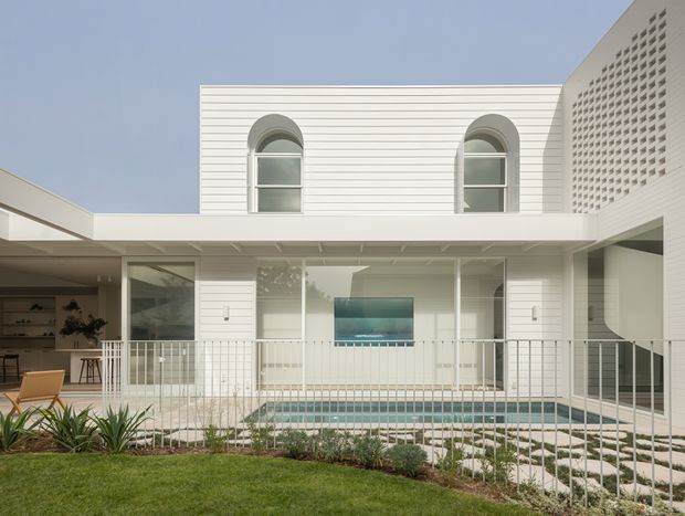 Exterior view of a bright white multi-level residential courtyard, featuring horizontal wall cladding, arched upper windows, a perforated breeze-block screen, extensive glazing, a small swimming pool, and a paved garden foreground.