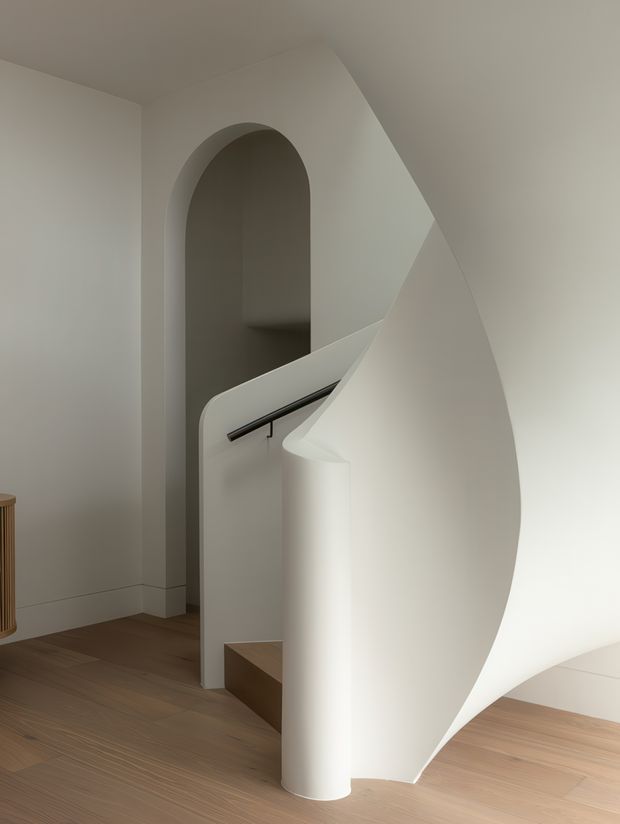 A sculptural curved staircase in a minimalist interior, featuring a smooth white plaster balustrade and sweeping spiral-like stair enclosure, dark metal handrail, pale timber flooring, and a tall arched wall opening. The restrained palette and sculptural geometry create a calm, refined atmosphere.
