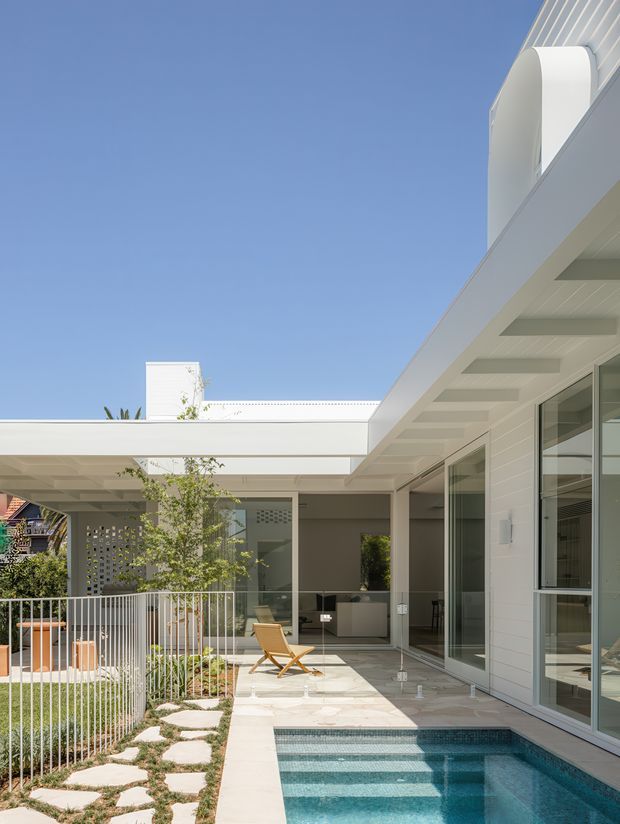 A bright contemporary courtyard exterior with white horizontal cladding, broad flat roof forms, deep eaves, extensive floor-to-ceiling glazing, and a compact tiled swimming pool. A paved terrace with a lounge chair borders landscaped planting, stepping stones, and a perforated privacy screen. The composition highlights the indoor-outdoor connection and crisp modern architectural detailing under a clear blue sky.