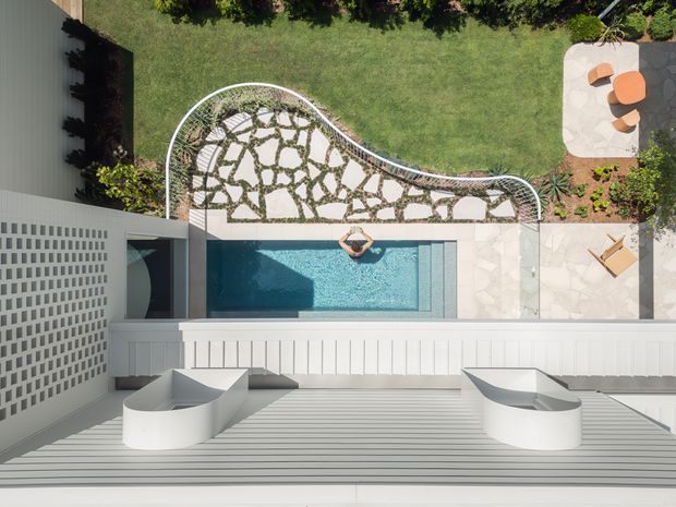 Aerial view of a contemporary residential courtyard with a narrow rectangular swimming pool, pale stone paving, a curved pebble-and-stepping-stone garden path, manicured lawn, outdoor seating, and white architectural structures with ribbed cladding and perforated breeze-block screening. The composition highlights the pool area, landscaped courtyard, and clean modern geometry.