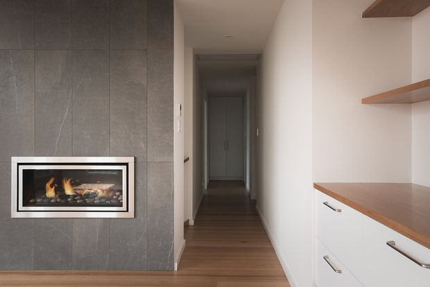 A contemporary residential hallway with a long timber floor leading toward a dark internal doorway. The left wall is clad in large-format dark grey tiles and contains a built-in linear fireplace with a white-trimmed surround. On the right, white built-in cabinetry with timber benchtops and open timber shelves provides storage. The space is narrow, clean-lined, and softly lit.