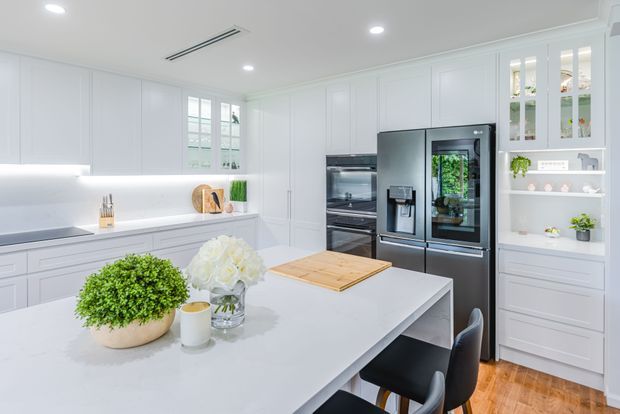 Bright, spacious white kitchen with extensive Shaker-style cabinetry, a large pale stone island, integrated stainless-steel appliances, glass-front display cabinets, open shelving, and warm timber flooring. Natural light enters through a window, while understated recessed lighting and concealed under-cabinet lighting create a clean, refined atmosphere.