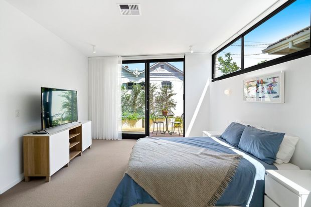 Bright contemporary bedroom with a neatly made bed, blue bedding, white walls, beige carpet, a low timber-and-white media cabinet with television, and expansive black-framed glazing. Vertical blinds open toward a small balcony or outdoor area, with neighbouring residential rooftops visible outside. The room has clean lines, abundant daylight, and understated furnishings.
