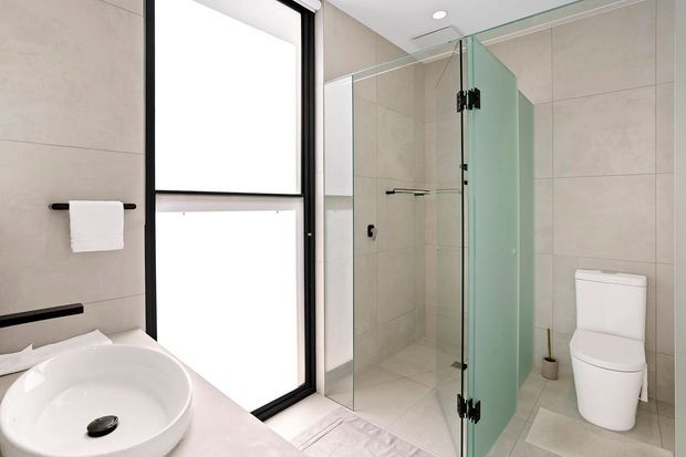 Bright contemporary bathroom with large-format beige wall and floor tiles, a white vessel basin on a pale benchtop, a frameless glass shower enclosure with black hardware, a white toilet, and a tall black-framed window. The space is clean, minimal, and softly lit by natural light and recessed ceiling lighting.