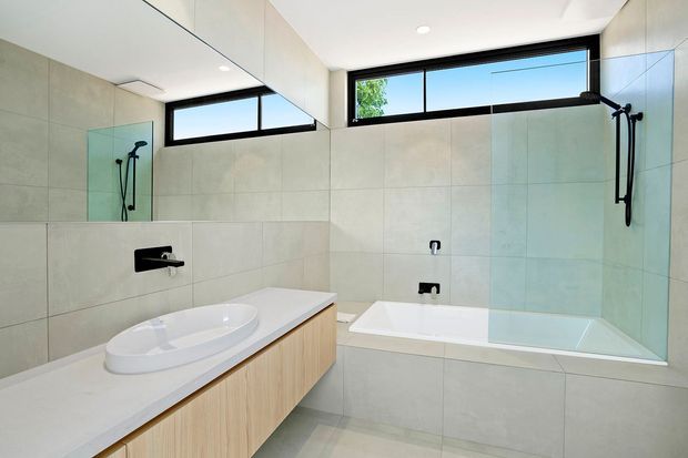 Bright contemporary bathroom with a freestanding-style white oval basin on a timber-look floating vanity, a built-in bathtub, large pale grey wall and floor tiles, black wall-mounted tapware and shower fittings, frameless glass shower screening, and high horizontal windows. The room has a clean, minimal, light-filled appearance.