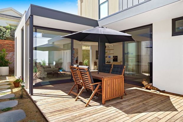 A bright contemporary courtyard-style exterior with a timber deck and outdoor dining setting beneath a large dark umbrella. White rendered walls, dark metal-framed floor-to-ceiling glazing, and a boxy flat-roofed form surround the deck. The glazing reveals a furnished interior living area and kitchen beyond, while planted edges and neighbouring residential fabric frame the space.