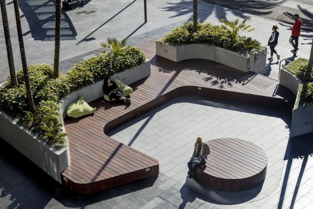 Elevated view of a landscaped urban terrace or courtyard with curved built-in timber decking and seating, large white planter beds filled with greenery and palm trees, and a central paved open area. The contemporary public or commercial outdoor space includes integrated benches and sculptural rounded forms.