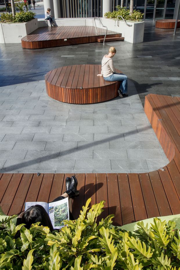 A landscaped urban plaza with broad grey stone paving, raised timber decking, and several curved, circular timber seating platforms. Integrated planters with lush greenery surround the space, while stainless-steel handrails lead to a slightly elevated deck. People are seated and reading, indicating a public outdoor gathering area.