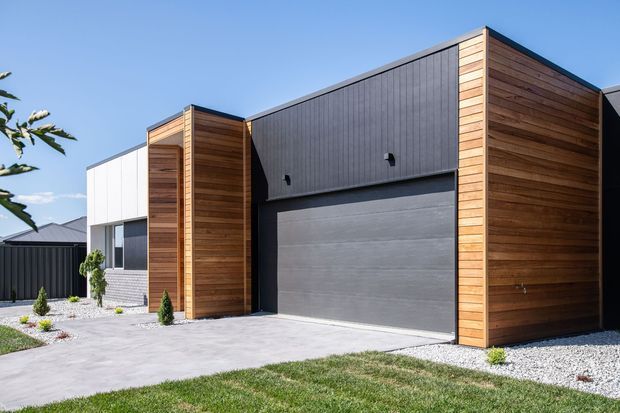 Contemporary single-storey residential facade with a prominent dark grey garage door, vertical dark cladding, and warm horizontal timber-clad volumes. The home is approached by a broad concrete driveway, with gravel landscaping and a neatly maintained lawn under clear daylight.