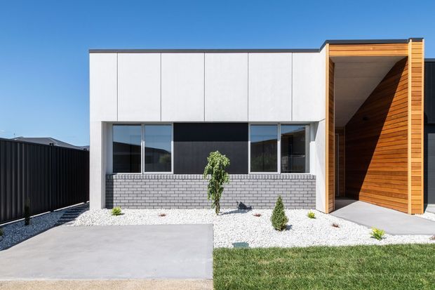 Front exterior of a contemporary single-storey house with a rectilinear white facade, dark-framed glazing, a grey brick lower wall, and a prominent timber-lined recessed entry or carport-like portal. The property has minimalist white gravel landscaping, a concrete driveway or path, lawn, and a dark boundary fence under a clear blue sky.
