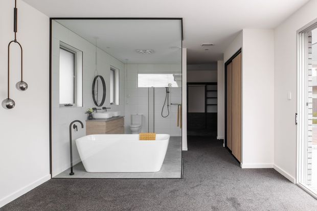 A bright contemporary bedroom with a freestanding white bathtub enclosed behind a large black-framed glass partition. The bathroom includes a wall-mounted vanity, toilet, shower, round mirror, and black tapware. Dark carpet extends through the bedroom, with built-in timber wardrobes and a large glazed door or window providing additional light.