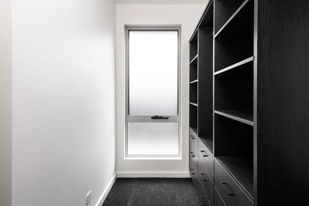 A narrow, minimalist built-in storage or wardrobe room with dark wood-grain cabinetry, open shelving, drawers, and a tall frosted-glass window. White walls and charcoal carpet create a clean, high-contrast interior.