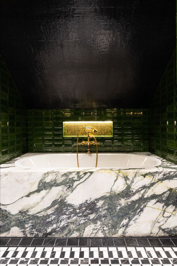A dramatic, highly stylised bathroom centred on a freestanding white bathtub. The walls are clad in glossy deep green rectangular tiles, with a small illuminated gold-toned tiled niche behind the tub and polished brass tapware. The bath front is wrapped in heavily veined white, grey and black marble, while the floor features a bold black-and-white checkerboard tile pattern. A dark textured ceiling creates a moody, luxurious atmosphere.