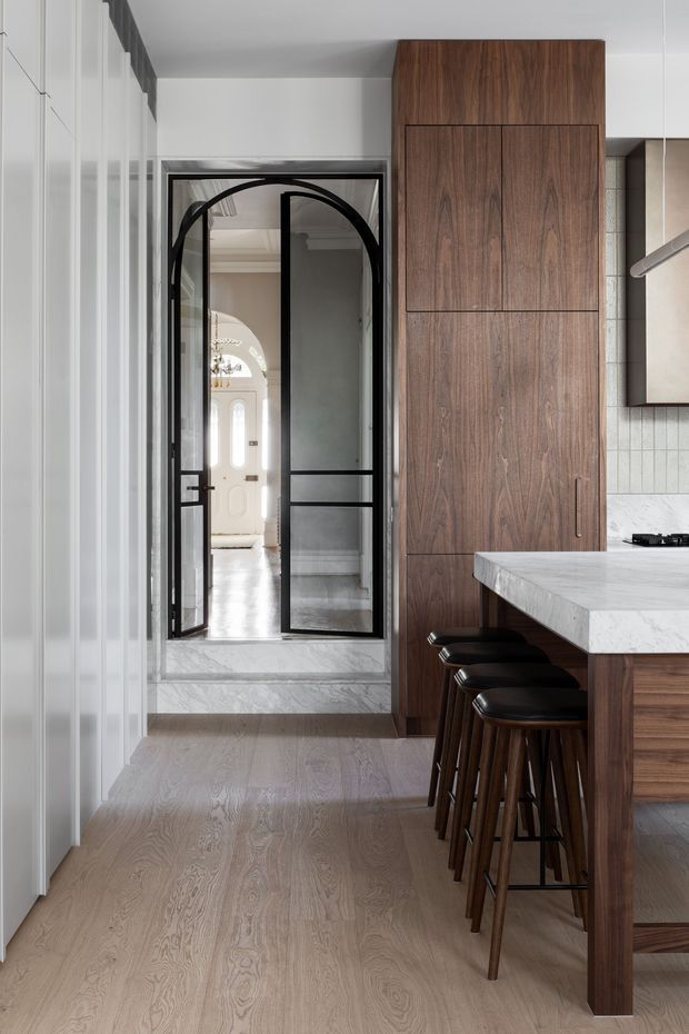 A refined kitchen interior viewed toward a black-framed glazed doorway. The space features a large natural wood kitchen cabinet and island, pale stone benchtop, white vertically detailed cabinetry, light timber flooring, and dark leather bar stools. The doorway opens to a brighter traditional hallway or entry with decorative architectural detailing.