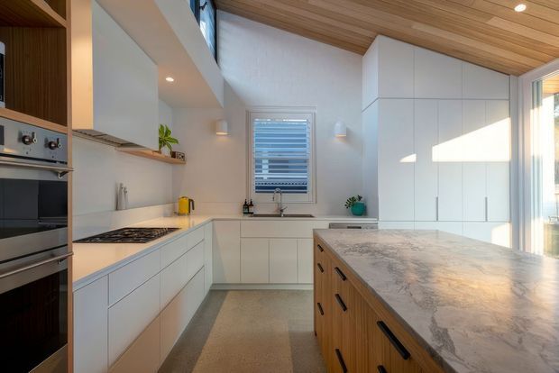 Bright contemporary kitchen with white flat-panel cabinetry, integrated appliances, a large timber-sided island with a pale stone benchtop, and a warm timber-lined ceiling. Natural light enters through the window and glazing, creating a clean, spacious, refined interior.