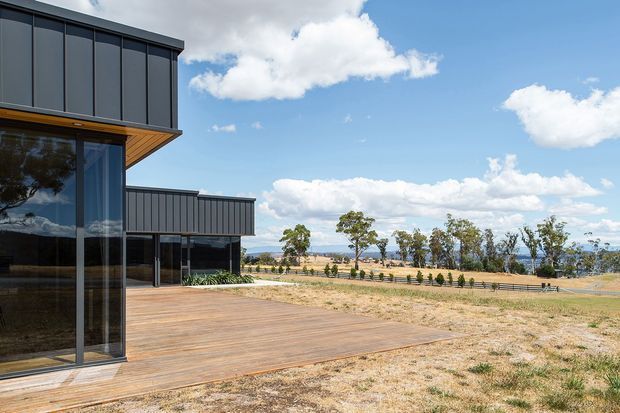 Contemporary multi-level building with dark vertical cladding, extensive floor-to-ceiling glazing and broad timber decking opening onto a dry rural landscape. The architecture has strong horizontal cantilevered rooflines and a clean rectilinear form, with distant water or low hills visible beyond the planted boundary.