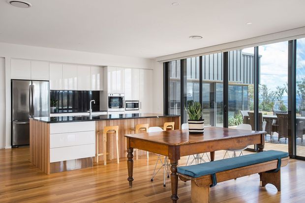 Bright, open-plan kitchen and dining area with extensive floor-to-ceiling glazing. The kitchen features white flat-panel cabinetry, a dark benchtop and splashback, integrated appliances, a stainless-steel refrigerator, and a timber island with bar stools. A substantial traditional timber dining table with a blue-cushioned bench and mixed dining chairs occupies the foreground. The glazing provides views toward water and surrounding landscape, while indoor plants and timber flooring add warmth.