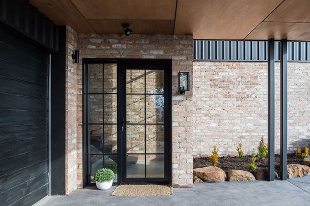 A covered residential entry porch with an industrial-style black framed glazed door set into an exposed pale brick wall. The porch has a warm plywood-lined ceiling, dark timber or painted cladding at the left, slim black steel posts, a concrete floor, and simple landscaped rock beds beyond. The glazing reveals a small interior entry area and stair beyond.