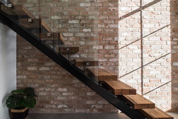 Interior view of a modern open-riser staircase with thick dark timber treads mounted against an exposed red brick wall. A black steel diagonal stringer and clear glass balustrade create an industrial, lightweight composition, with strong sunlight casting linear shadows across the masonry. A potted plant sits at the base of the stair.