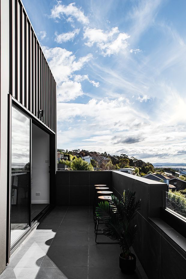 A contemporary upper-level terrace or balcony beside a modern building, with dark tiled flooring, black privacy walls and balustrades, slim black-framed glazing, and a row of bar stools along a narrow counter. The terrace overlooks a leafy suburban hillside and distant coastal or water views beneath a bright, partly cloudy sky.