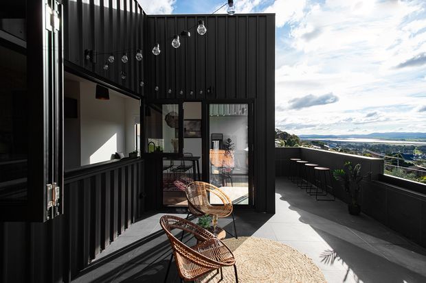 A sophisticated rooftop terrace or balcony with dark vertical metal cladding, an outdoor bar counter with stools, a small seating area, hanging string lights, potted plants, and a broad urban/coastal outlook. Large glazed doors connect the terrace to an interior space. The design has a dramatic, contemporary feel with strong black architectural surfaces and warm wicker furniture.