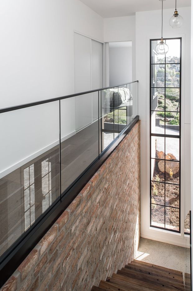 Bright contemporary stairway and upper landing with a black-framed glass balustrade, exposed reclaimed brick stairwell wall, dark timber stair treads, and a tall industrial-style window overlooking landscaped grounds. A bedroom is partially visible through the opening at the upper level. Clear pendant lights hang beside the window, while strong daylight emphasizes the double-height vertical space.