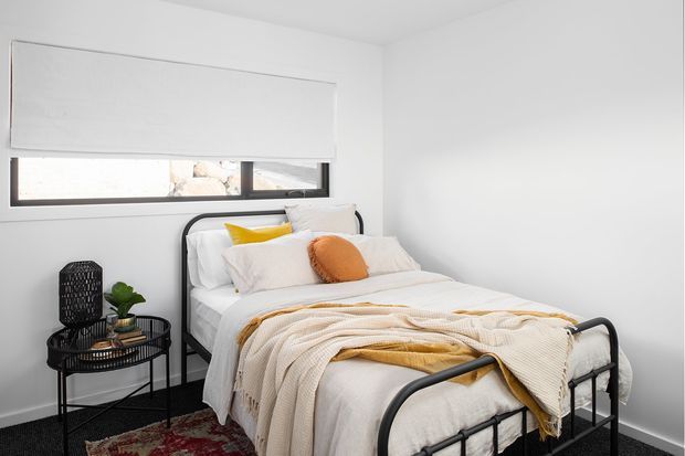 A compact, bright bedroom with a white upholstered bed dressed in layered cream and white bedding, mustard-yellow and rust-orange accent cushions, and a dark metal bed frame. A black round side table with a small plant sits beside the bed, while a wide horizontal window with a white roller blind provides natural light. The room has white walls, dark flooring, and a patterned rug.