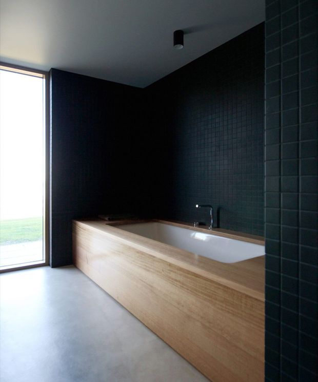 A dark, minimalist bathroom featuring a built-in freestanding-style bathtub enclosed in light timber paneling, a black wall-mounted mixer tap, and dark charcoal small-format tiled walls. A large glazed opening brings in daylight and views of grass, while the smooth pale floor and white ceiling provide contrast.