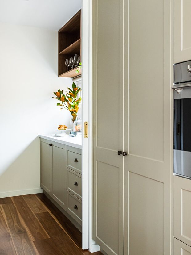 A bright, narrow kitchen pantry or butler's pantry viewed through an open doorway. The space features pale painted built-in cabinetry with dark knobs, a white benchtop, warm dark timber flooring, open dark-wood shelving displaying glassware, and a small arrangement of foliage. Tall matching cabinetry and integrated appliances are visible at the right edge.