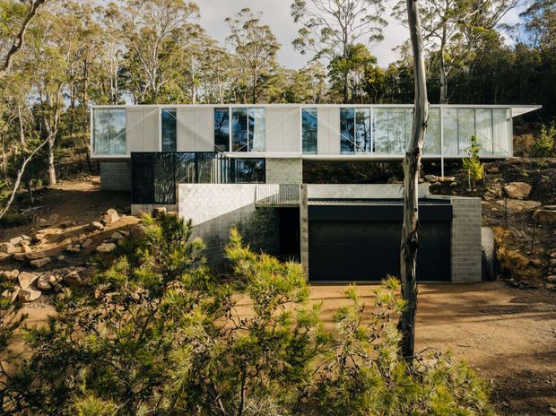Exterior view of a contemporary elevated residence set into a wooded, sloping site. The long upper level features extensive glazed walls with pale framing and projecting geometric volumes, while the lower level is formed in dark concrete or masonry and contains a large garage opening. Native bushland and rock landscaping surround the house.