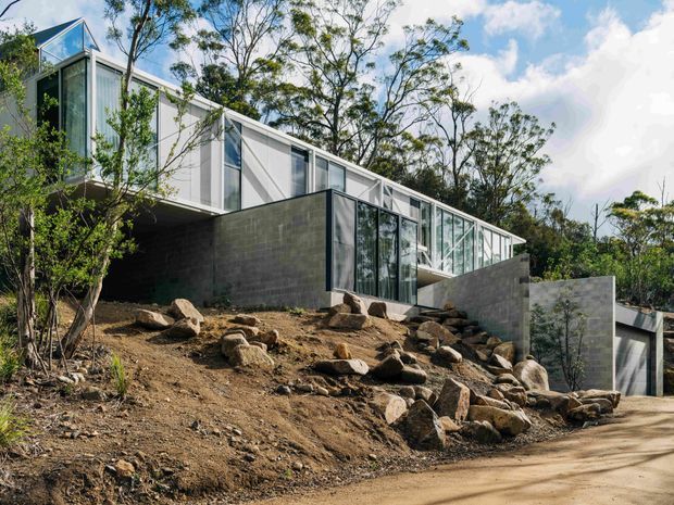 Exterior view of a contemporary multi-level home set into a steep, rocky, tree-covered site. The building combines extensive floor-to-ceiling glazing, light vertical cladding, dark concrete block walls, and projecting volumes, with a long glazed upper level following the slope.