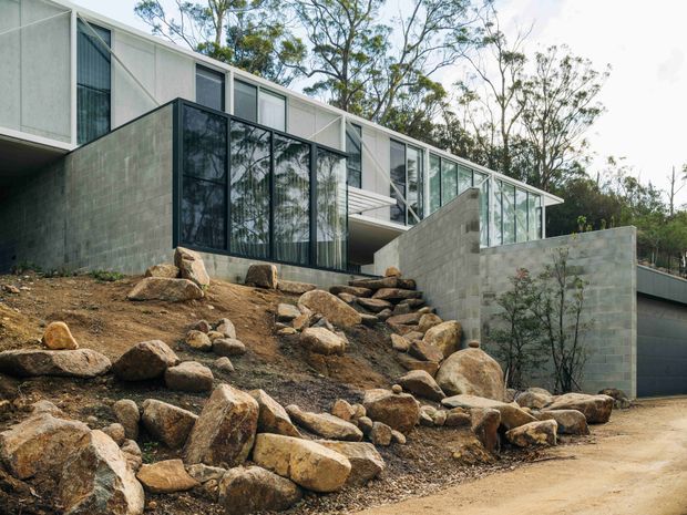 Exterior view of a contemporary multi-level house set into a rocky, sloping site. The building combines pale concrete block walls, extensive floor-to-ceiling glazing with dark frames, and a long low-pitched roofline, with a garage visible at the lower right. Native trees and exposed boulders surround the structure.