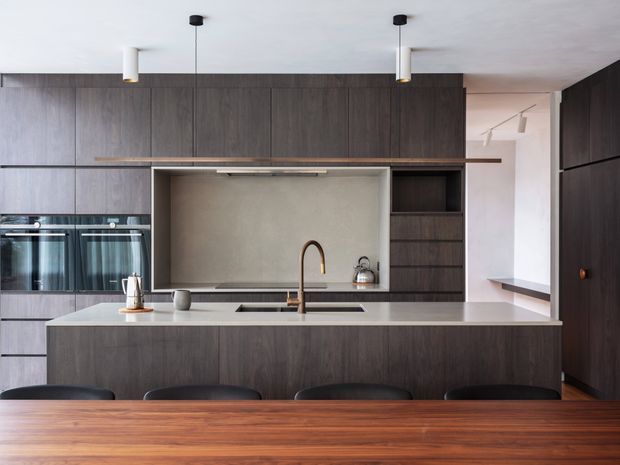 Contemporary kitchen with a large dark timber island and extensive full-height cabinetry. The island includes an integrated sink and warm metallic tap, while a pale stone benchtop contrasts with the dark wood joinery. Built-in ovens, a recessed cooking area with a neutral splashback, pendant lights, and a timber dining table with dark chairs are visible.