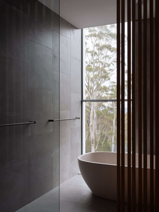 A moody, contemporary bathroom with large dark grey wall and floor tiles, a frameless glass shower, slim horizontal towel rails, and a sculptural freestanding white bathtub positioned beside a full-height window. Vertical dark timber screening partially separates the bath area, with eucalyptus trees visible outside.