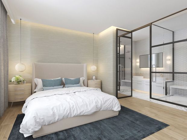 A bright, refined contemporary bedroom with a large upholstered bed, pale textured wall finish, light timber flooring, and a dark area rug. Floor-to-ceiling black-framed glass partitions reveal a modern bathroom with a freestanding bath, vanity, and shower area. Soft globe pendants and concealed cove lighting create a calm, luxurious atmosphere.