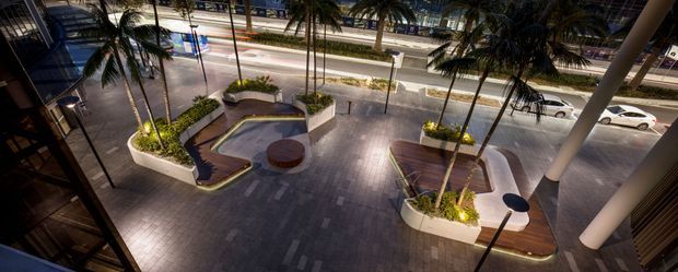 Elevated nighttime view of a landscaped paved forecourt or terrace beside a multi-lane urban street. Large palm trees sit in raised curved planters with integrated warm lighting, benches, and small round tables, creating a contemporary outdoor gathering space beneath a canopy or building overhang. Commercial buildings, illuminated storefronts, and passing vehicles are visible beyond.