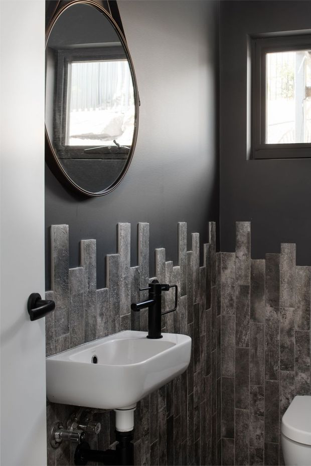 Compact powder room with a wall-mounted white ceramic basin, matte black tapware and plumbing, and a partially visible toilet. Dark charcoal painted walls contrast with a dramatic vertical arrangement of textured grey tiles around the lower walls. A large oval framed mirror and small windows bring reflected and natural light into the space.