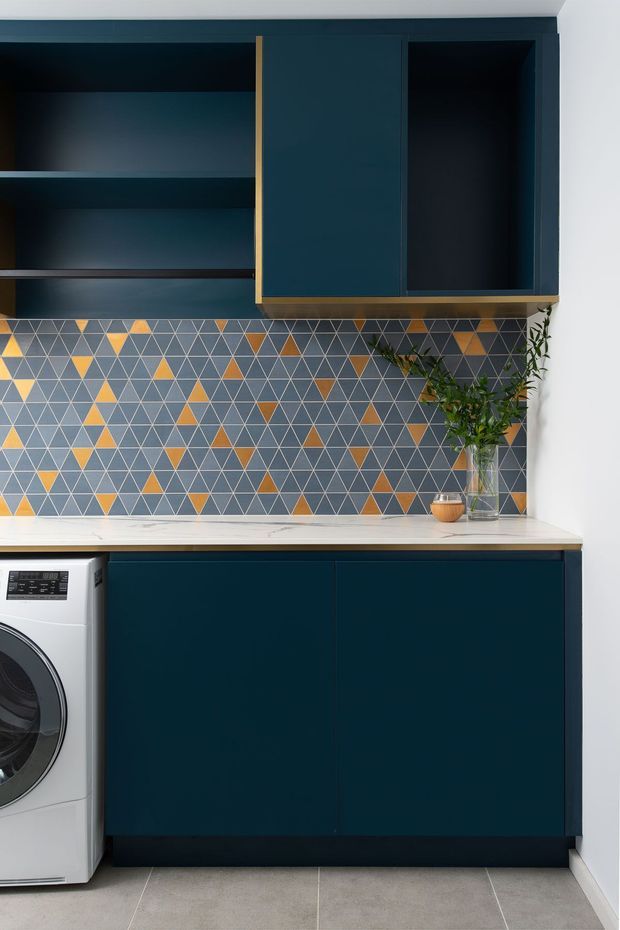 A compact, contemporary laundry room with dark navy built-in cabinetry, open shelving, and brass-toned trim. A geometric patterned tile splashback in grey, white, mustard, and terracotta sits above a pale stone-look benchtop. A front-loading washing machine is partially visible at left, while a glass vase with leafy branches adds a natural accent. Large grey floor tiles and white walls complete the space.