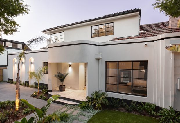 Exterior view of a refined two-storey residence with white rendered walls, dark tiled pitched roofs, black-framed gridded windows, a curved upper-level facade, covered entry, and landscaped front garden with a paved walkway. Warm exterior lighting highlights the contemporary-traditional facade.