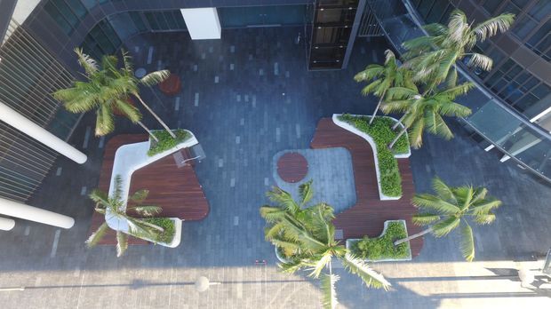 Aerial view of a contemporary commercial atrium or courtyard with dark tiled paving, curved timber deck-like seating platforms, white raised planters, and several palm trees. Glass façades, louvres, and surrounding upper-level circulation frame the landscaped internal space.