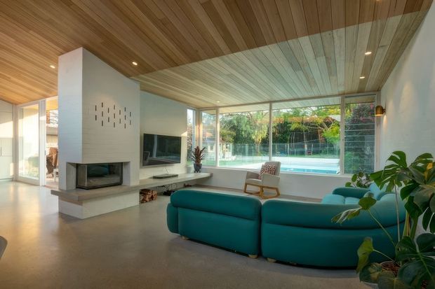 A bright, open-plan living room with a large teal sectional sofa facing a television and a sculptural double-sided fireplace. Floor-to-ceiling glazing looks onto a swimming pool and landscaped garden. The room features extensive warm timber ceiling lining, pale walls, polished concrete flooring, and abundant indoor planting.