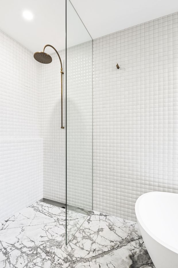Bright contemporary bathroom featuring a frameless glass shower enclosure, white small-format tiled walls, a sculptural freestanding white bathtub, and dramatic white marble-look flooring with dark grey veining. A bronze rainfall shower and fittings provide a warm metallic accent.