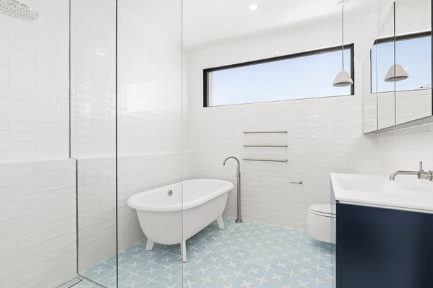 Bright contemporary bathroom featuring white tiled walls, a freestanding white bathtub, frameless glass shower enclosure, wall-mounted vanity with dark blue cabinetry, chrome fixtures, and a patterned pale blue and white floor. High horizontal glazing and pendant lights add a clean, airy feel.