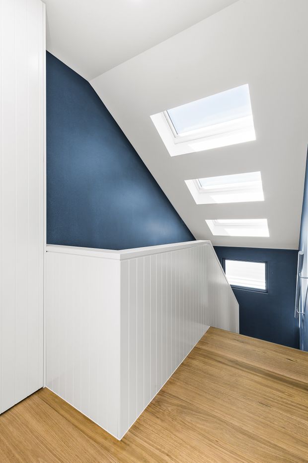 Bright contemporary stairway with warm timber flooring, white vertical-panelled stair balustrade and cabinetry, deep navy-blue walls, and multiple skylights set into a sloping ceiling. A small window illuminates the lower stair landing.