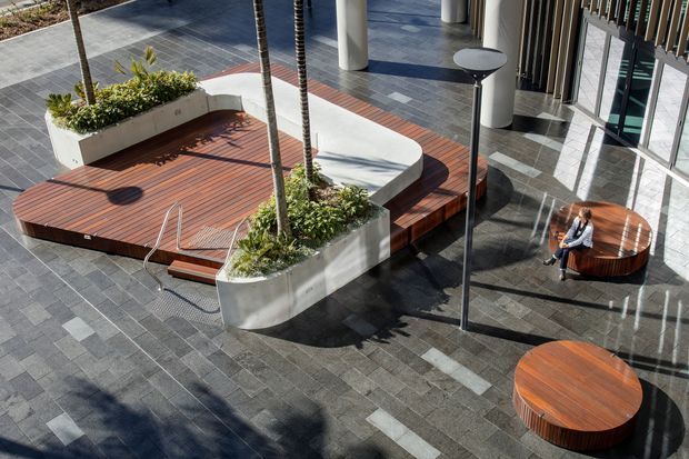 A contemporary indoor-outdoor communal courtyard or atrium with dark stone paving, raised timber platforms, integrated white planters, palm trees, curved concrete seating, and round timber tables. Floor-to-ceiling glazing and vertical fins line the building edge, while a person sits at one of the tables.