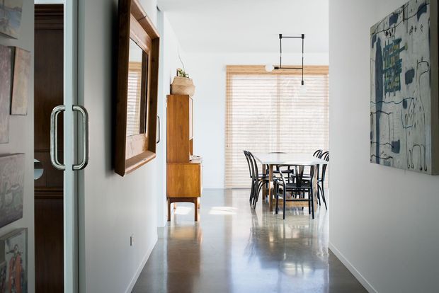 View through a glazed entry or hallway toward a bright dining area. The space features polished concrete flooring, white walls, timber-framed glazing with horizontal blinds, a timber sideboard, framed artwork, and a black dining table with matching chairs beneath a simple linear pendant light.