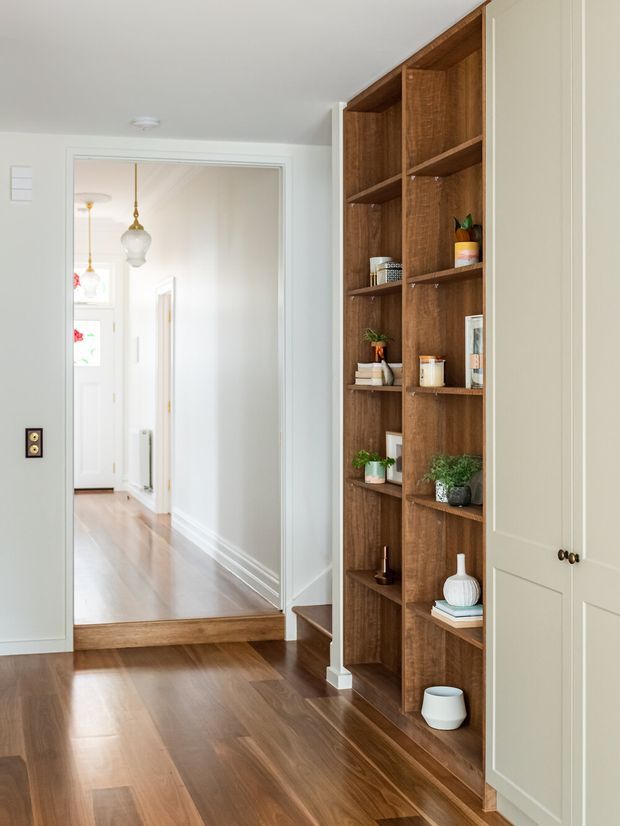 Bright residential hallway with polished dark timber flooring, white walls and trim, and a full-height built-in timber display shelving unit beside pale built-in cabinetry. A large opening leads to a raised, light-filled corridor with a decorative pendant and additional white doors. The space is tidy and refined, with warm wood joinery and small decorative objects on the shelves.