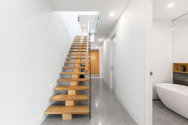Bright minimalist interior hallway with an open-riser timber staircase, clear glass balustrade, polished grey terrazzo flooring, white walls and recessed ceiling lighting. A partially visible contemporary bathroom with a freestanding white bathtub and timber feature shelving is seen through the doorway on the right.